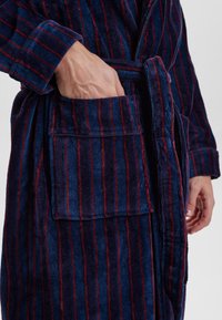 Accappatoio a righe texture navy e rosso con cintura e tasca frontale. Materiale morbido e peloso; polsini e colletto elegantemente rifiniti.