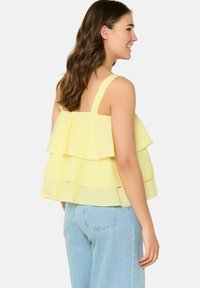 Rumena karirasta bluzica z širokimi naramnicami, ki ima tri plasti volančkov in bolj ohlapen kroj, v kombinaciji z svetlo modrimi kavbojkami.