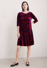 Röd sammetklänning med blå blomstermönster, trekvartsärmar och rund halsringning; matchad med svarta, lackiga Mary Jane-skor.