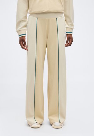 Μπεζ παντελόνι με φαρδιά μπατζάκια και κάθετη σκούρα πράσινη ρίγα στο μπροστινό μέρος, συνδυασμένο με μπεζ τοπ και ανοιχτόχρωμα αθλητικά παπούτσια.