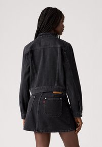 Chaqueta de mezclilla negra con silueta recortada, mangas largas y puños abotonados. Combinada con una falda de mezclilla negra a juego, que presenta bolsillos y detalles de costura.