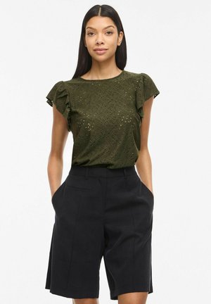 Žena dugom tamnom kosom nosi maslinasto zelenu bluzu s izrezima i naboranim rukavima te crne široke hlače visokog struka, ruke u džepovima.