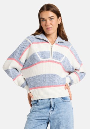 Vrouw draagt een blauw-wit-roze gestreepte trui met een halve ritssluiting aan de kraag, gecombineerd met lichtblauwe spijkerbroek, handen in de heupen.