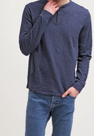 Person, der ein langärmeliges navyblaues Henley-Shirt und eine blaue Jeans trägt, eine Hand in der Nähe des Kinns und die andere in der Tasche.