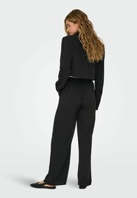 Veste noire raccourcie à manches longues, associée à un pantalon noir ample. Tissu lisse et design décontracté. Chaussures plates noires.