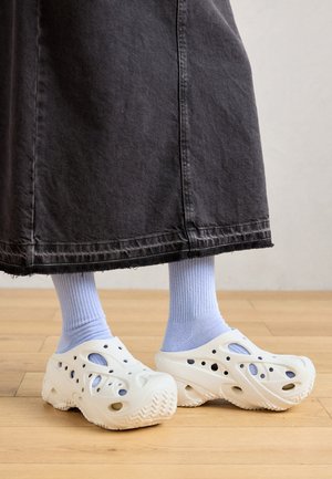 Voeten in lichtblauwe sokken en off-white geperforeerde instapschoenen, met de zoom van een zwarte rok zichtbaar boven een houten vloer.