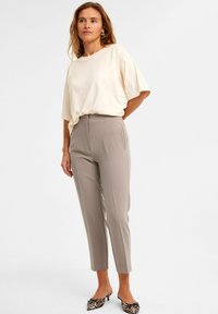 Beige ylin kokoinen t-paita lyhyillä hihoilla, yhdistettynä harmaisiin muotoiltuihin housuihin, joissa on sivutaskut; mallilla on päällään eläinkuosilla varustetut korkokengät.