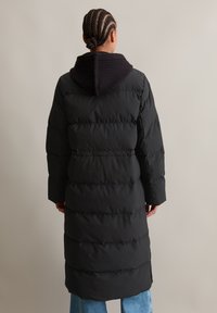 Persoon met gevlochten haar, gekleed in een lange zwarte pufferjack met een geribde capuchon, staand tegen een effen lichte achtergrond.