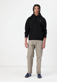 Schwarzer Baumwollhoodie mit einer Fronttasche und Kordelzug; kombiniert mit hellbeigen Hosen und blauen Schuhen mit weißen Streifen.