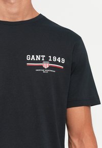 Sort t-shirt med korte ærmer, hvid "GANT 1949" tekst, rød og hvid stribet skildemblem, og "American Sportswear NH.CT." under.