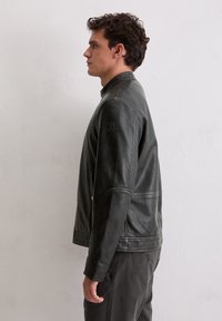 Junger Mann in einer dunkelgrauen Lederjacke und passenden Hosen steht im Profil vor einer schlichten hellen Wand.