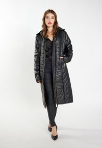 Lang, sort, quilted pufferjakke med hætte, lynlåslukning foran og sidelommer. Bæres over en sort top og tætsiddende bukser. Glat, skinnende tekstur.