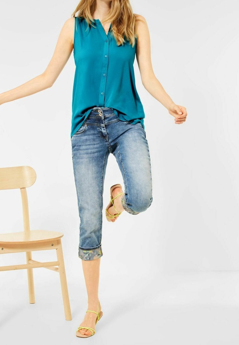 Blouse sans manches turquoise, jean en denim bleu clair coupé avec des ourlets retroussés et sandales beiges avec des brides vertes. Chaise en bois en arrière-plan.