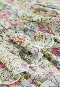 Tissu avec imprimé floral multicolore en rose, jaune, vert et violet, avec des détails en dentelle œillet blanche brodée et des plis doux.