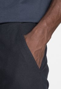 Right hand of a person with dark skin inserted into the pocket of dark blue pants, wearing a navy blue shirt.
