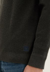 Suéter de punto marrón oscuro con puños acanalados. Presenta una pequeña etiqueta cuadrada en el dobladillo con un logotipo. Textura suave, diseño casual.