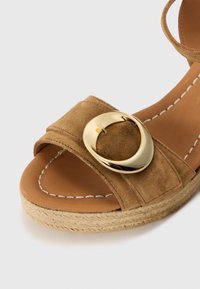 Sandalo in suede beige con una larga fascia e una grande fibbia circolare dorata, dotato di una suola a cuneo avvolta in juta e cuciture bianche in contrasto.