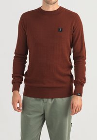 Maglione lavorato a maglia color ruggine con un motivo testurizzato, scollatura rotonda e polsini a costine. Presenta un piccolo ricamo del logo nero sul petto.