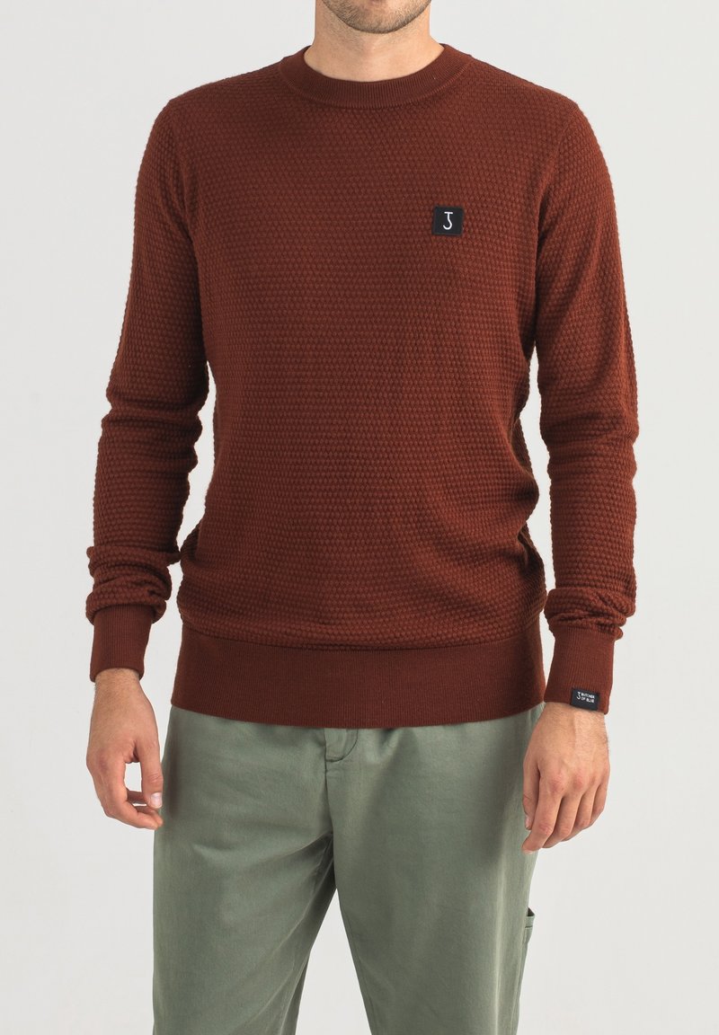 Maglione lavorato a maglia color ruggine con un motivo testurizzato, scollatura rotonda e polsini a costine. Presenta un piccolo ricamo del logo nero sul petto.