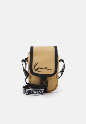 Petit sac bandoulière beige avec bordure noire, rabat avant, boucle noire et sangle ajustable avec motif de texte blanc.