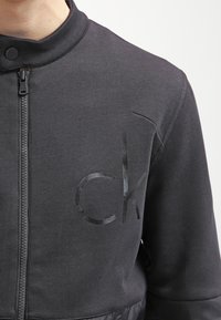 Schwarze Zip-Jacke aus strukturiertem Stoff, mit hohem Kragen und glänzendem "ck"-Logo auf der Vorderseite. Doppelte Reißverschluss-Details.