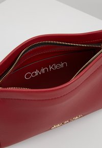 Czerwona skórzana torebka Calvin Klein z złotym zamkiem błyskawicznym, z widocznym logo marki w otwartej głównej komorze.