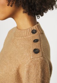 Beige Wollpullover mit einem gerippten Kragen und drei dunklen Knöpfen an der Schulter. Weicher Stoff mit einem klaren, minimalistischen Design.