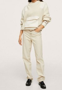 Beżowy sweter na drutach z dużą przednią kieszenią i podwiniętymi mankietami, zestawiony z jasnobeżowymi spodniami o prostych nogawkach i czarnymi mokasynami.