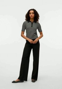 Mulher com cabelo encaracolado, vestindo uma blusa de manga curta listrada em preto e branco, calças largas pretas e sapatos estampados, contra um fundo branco.