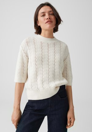 Junge Frau mit kurzen braunen Haaren trägt einen weißen gestrickten Kurzarmpullover und dunkelblaue Jeans vor einem schlichten hellen Hintergrund.