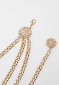 Accessorio in catena d'oro con un elegante fermo circolare decorato da un motivo ornamentale e tre catene collegate, presentando una finitura lucida.