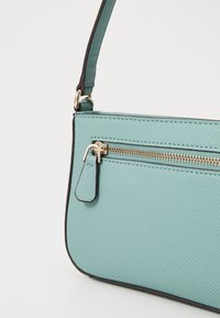 Sac à main turquoise en matériau texturé avec une poche zippée, des bordures marron et une bandoulière réglable. Détail de quincaillerie discret près de la fermeture éclair.