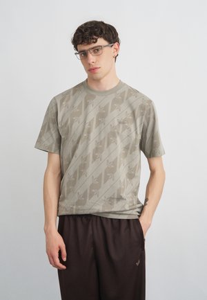 Junger Mann mit lockigem dunklem Haar und Brille, der ein gemustertes beiges T-Shirt und dunkelbraune Hose trägt, steht vor einem einfarbigen hellen Hintergrund.