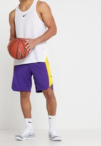 Valkoinen Nike-toppi, purppuraiset shortsit keltaisilla sivuakkenteilla, teksturoitu kangas, harmaat Nike-tennarit, pitelee koripalloa.