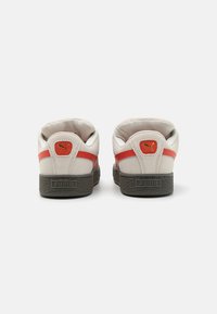 Sneaker bianche con accenti rossi, realizzate in suede e materiale in pelle, design low-top, suola grigia texturizzata, con logo Puma sul tallone.