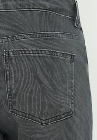 Los pantalones vaqueros gris oscuro presentan un patrón texturizado con líneas onduladas. El bolsillo trasero es visible, mostrando su elegante costura y estructura.
