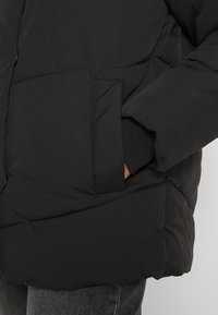 Personne portant une veste noire matelassée avec la main placée dans la poche avant, un poignet visible et un pantalon gris sur un fond neutre.