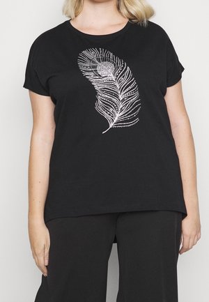 Femme portant un t-shirt noir à manches courtes avec un motif détaillé de plume de paon blanche sur le devant, associé à un pantalon noir.