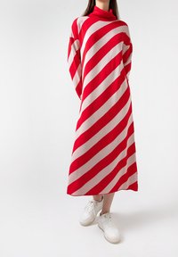 Personne portant une robe longue rayée rouge et blanche en diagonale avec un col montant, associée à des baskets blanches sur un fond blanc.