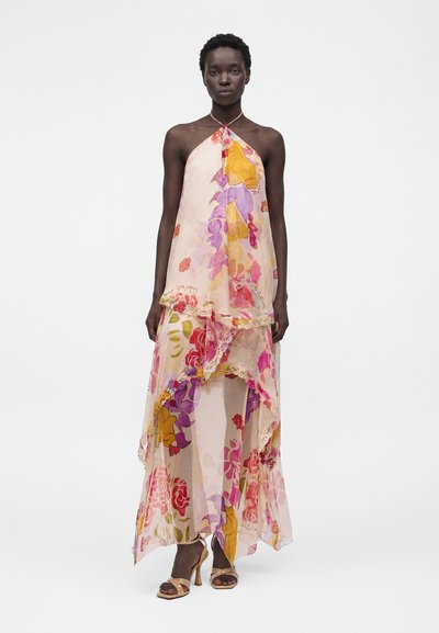 Ψηλή γυναίκα φοράει ένα ρέον, διαφανές φόρεμα halter με μεγάλα πολύχρωμα λουλουδάτα σχέδια και στρωμένα κυματιστά τελειώματα, συνδυασμένο με μπεζ σανδάλια με ψηλό τακούνι.