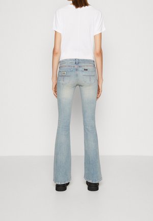 Kobieta ubrana w jasnoniebieskie jeansy z rozszerzanymi nogawkami i białą koszulkę z krótkim rękawem, stoi z tyłu na jednolitym białym tle.