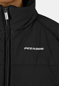 Svart polstret jakke med høy krage, glidelås foran, og ordet "PEGADOR" i hvitt på venstre brystområde. Glatt tekstur og slank design.