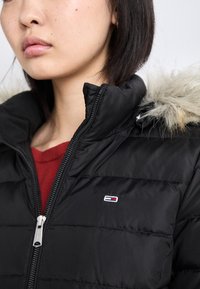 Mustpehme sulejope karvase kapuutsiga, ees lukk ja rinnal logo, kihiliselt punase pluusi peal. Sile tekstuur.