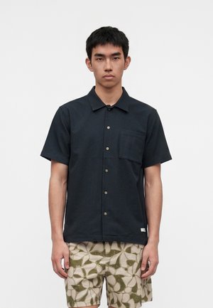 Jeune homme portant une chemise noire à manches courtes boutonnée et un short beige avec des motifs abstraits de feuilles vertes, debout devant un fond uni.