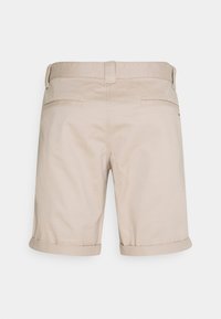 Short beige pour hommes avec poches passepoilées à l'arrière, passants pour ceinture et ourlets retroussés, vus de dos sur un fond uni.