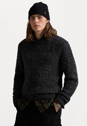 Jonge man met lang haar, die een zwarte gebreide beanie draagt, een gestructureerde donkergrijze trui over een groen geruit overhemd, zwarte broek en een horloge.