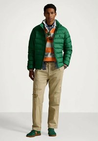 Muškarac u zelenoj puffer jakni, narančastom i sivom prugastom puloveru, bež izrezanim hlačama i zelenim tenisicama, stoji naslonjen o jednoličnu pozadinu.