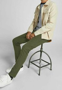 Giacca beige, camicia a righe blu navy e bianca, pantaloni verde oliva e sneakers bianchi. Seduto su uno sgabello di metallo. Sfondo neutro.