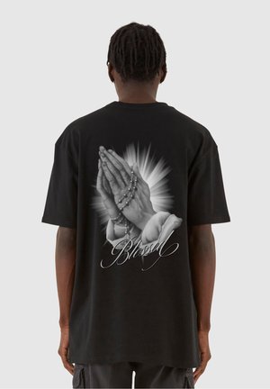 Schwarzes oversized T-Shirt mit einer Grafik von betenden Händen mit Rosenkranzperlen und dem Wort "Blessed" in weißer Schreibschrift auf der Rückseite.