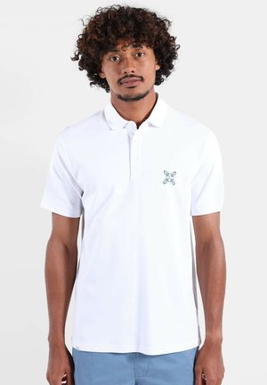 Homme aux cheveux bouclés portant un polo blanc à manches courtes avec un petit logo à motif sur la poitrine et un pantalon bleu, sur un fond uni.